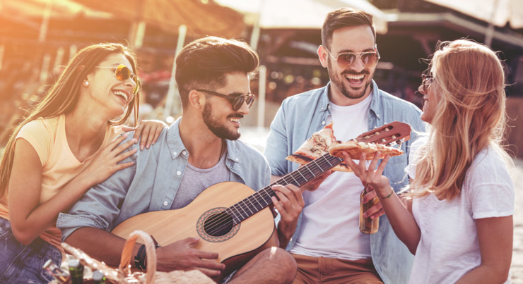 a group of young adults around a man playing guitar and everyone eating pizza